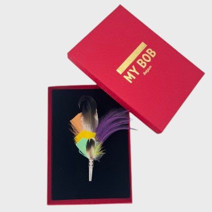 Purple, yellow, light pink and green, and black feather brooch in a box on a light gray background