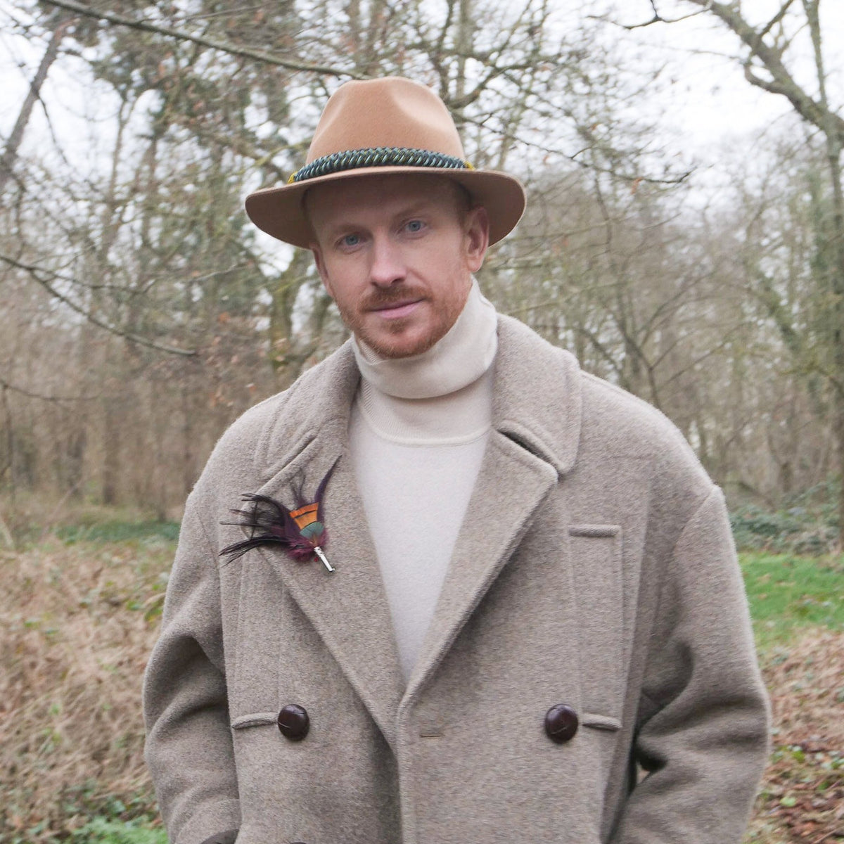 A person wearing a beige coat adorned with a purple and orange dyed feather brooch.