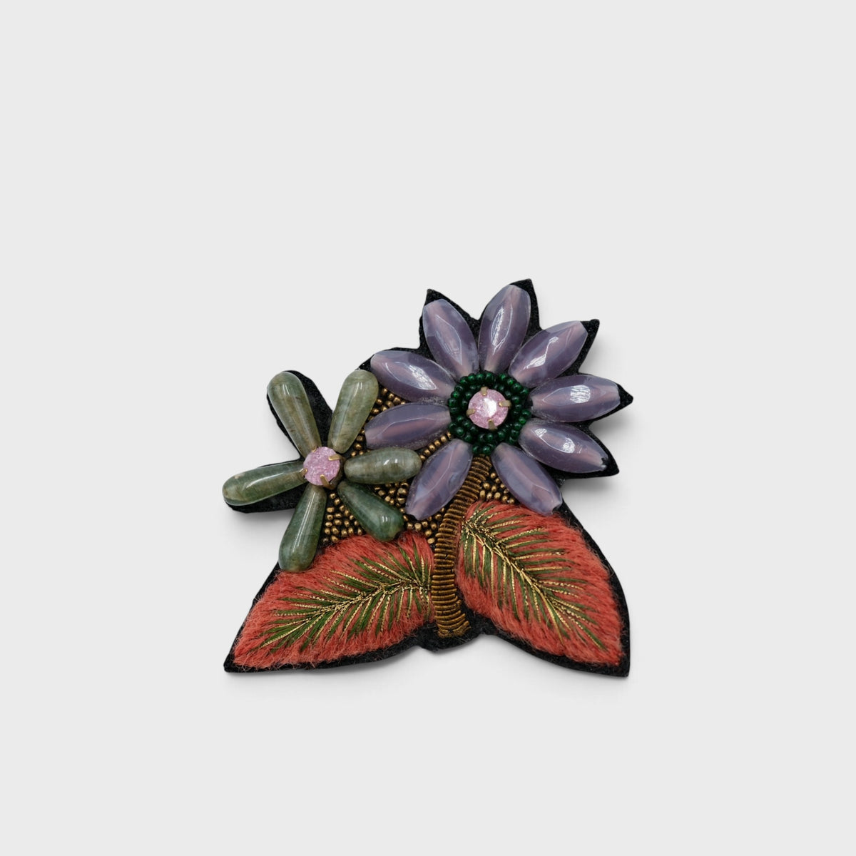 A floral brooch featuring two stylized flowers and leaves. The larger flower on the right is lavender-purple with a dark green center and a pink gem accent.