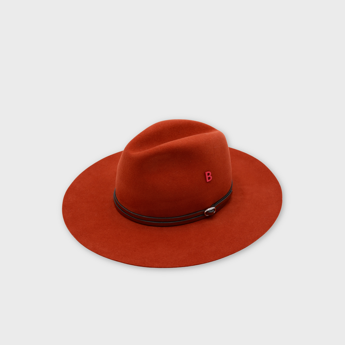 A rust-orange felt wide-brim hat with a dark brown leather band and a small red letter 'B' pin on the side, shown on a light gray background.