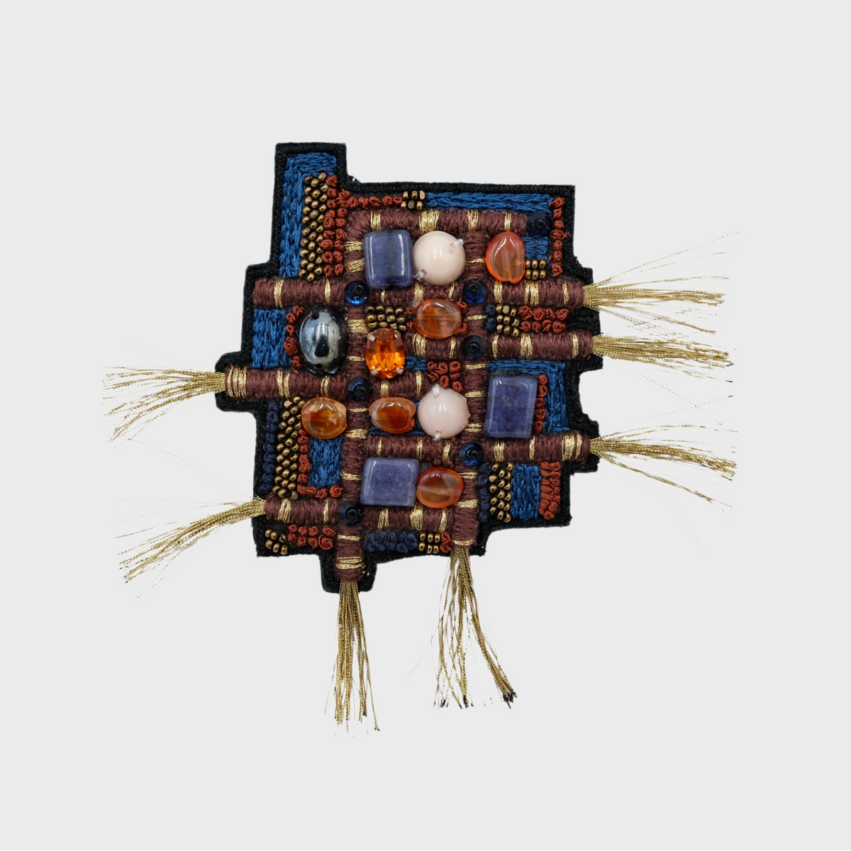 A square, handcrafted beaded and woven brooch featuring geometric patterns and stones in blue, red, brown, white, and black, with small golden frayed threads extending from the sides, shown against a plain light gray background.