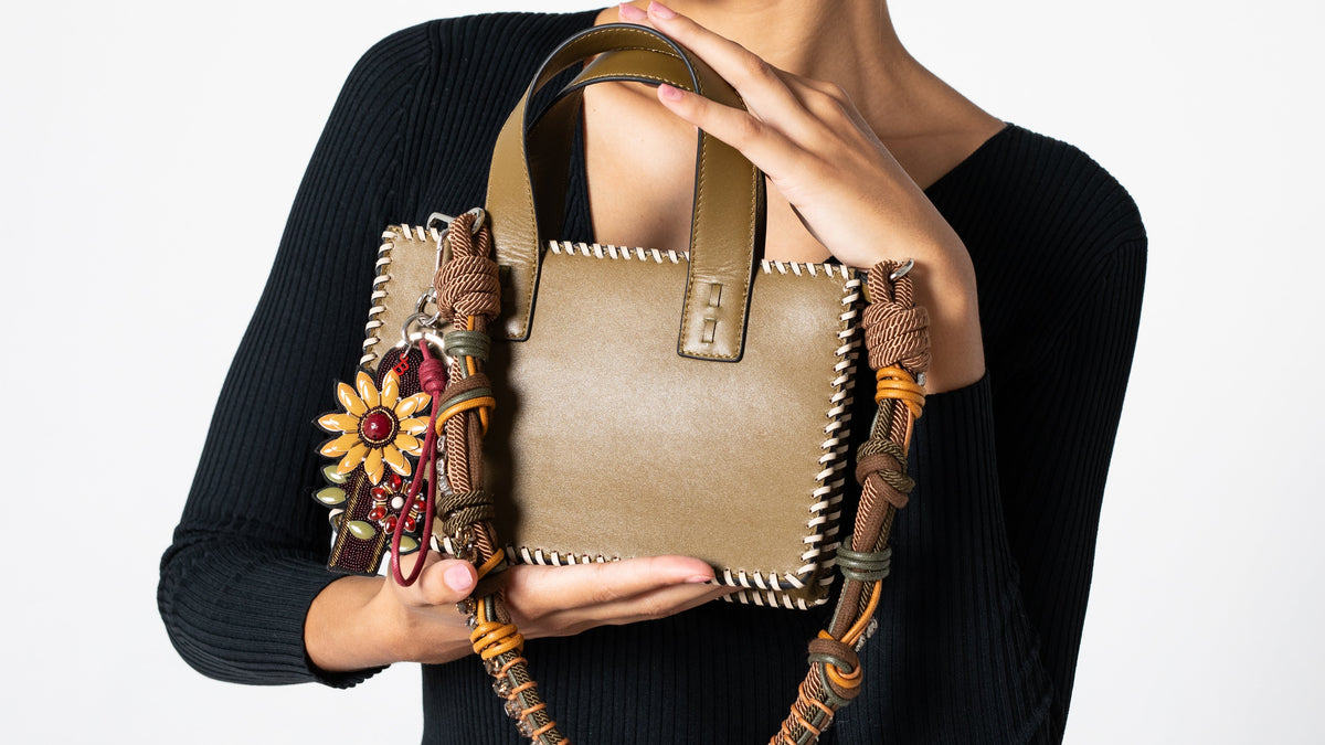 Person holding a decorative handbag with an embroidered bag charm with red details and two flowers on a plain background