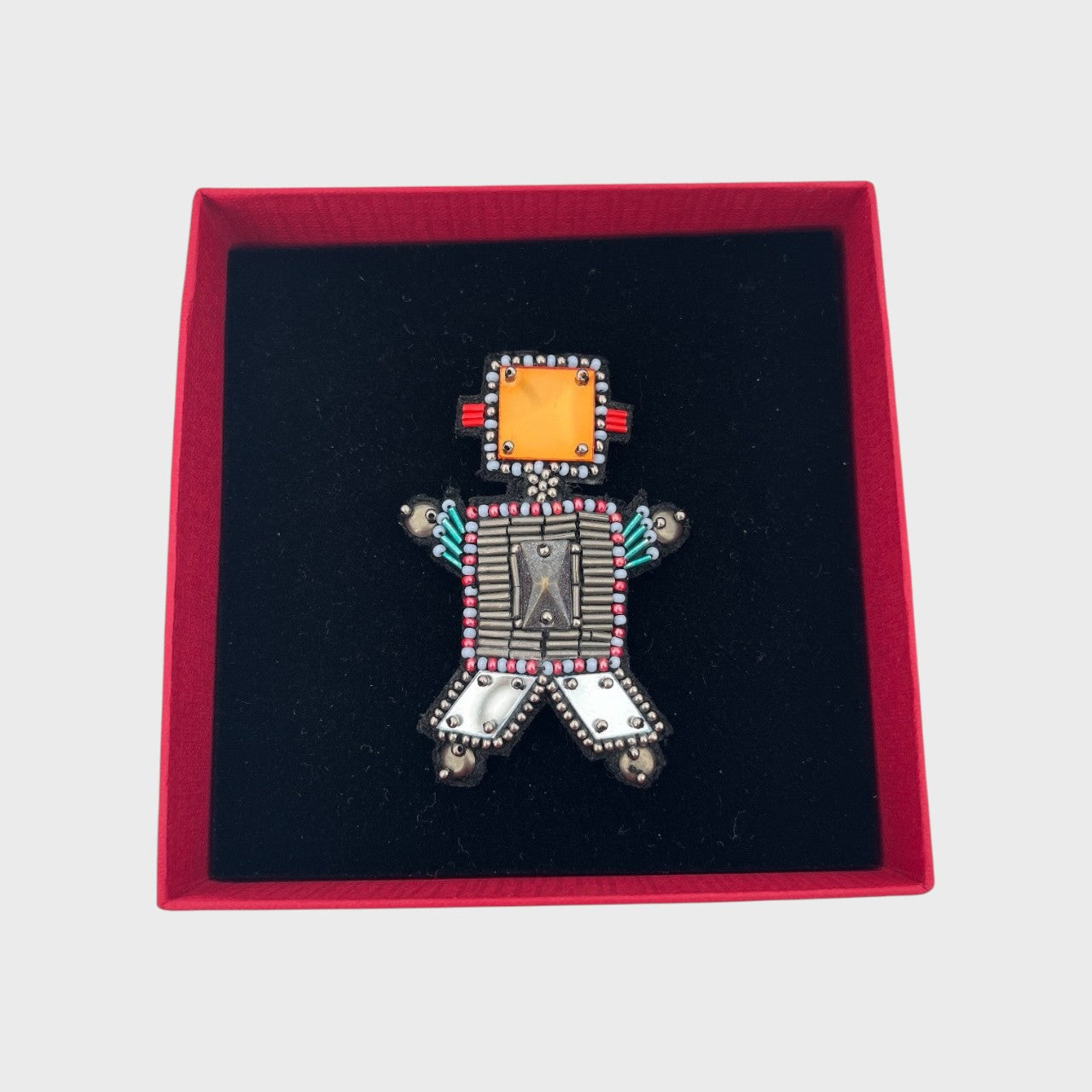 An embroidered brooch representing a grey robot with an orange head in a box. 