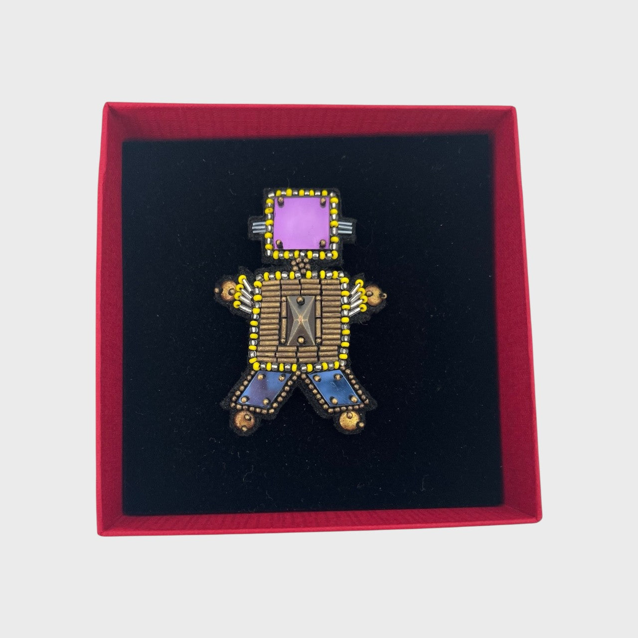 An embroidered brooch representing a bronze robot with a purple head in a box. 