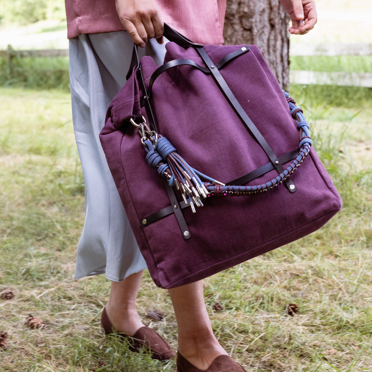 Person holding a purple optimiste tote bag with decorative bag charms and a colourful macrame shoulder strap in an outdoor setting.
