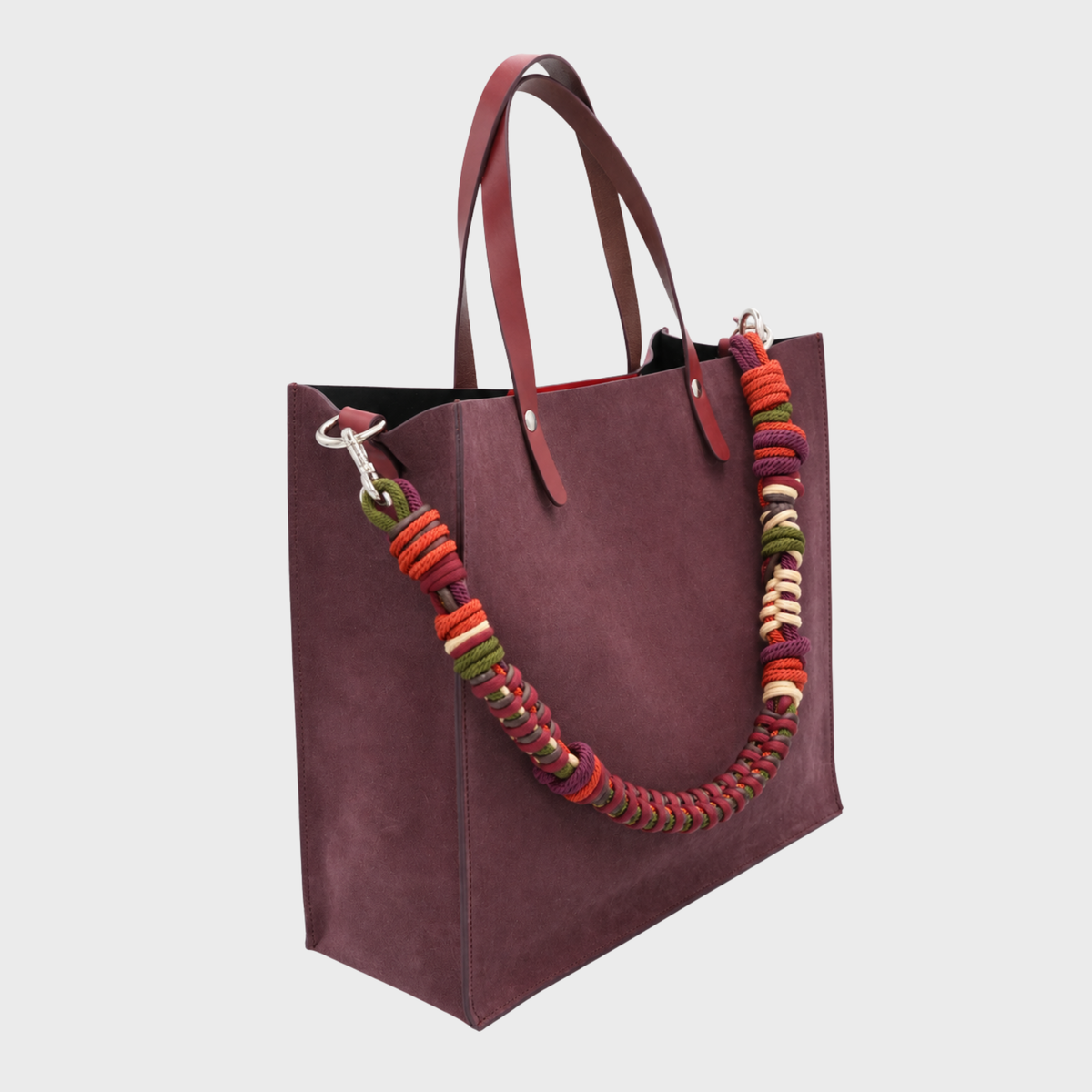 A large structured tote bag in deep burgundy suede, featuring dual leather handles and a thick, colorful braided shoulder macrame strap made in red, orange, green, and cream, shown against a light gray background.