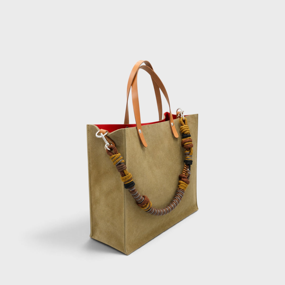 A large green suede tote bag with light brown leather shoulder straps and a thick braided multicolor macrame strap, shown against a light gray background.