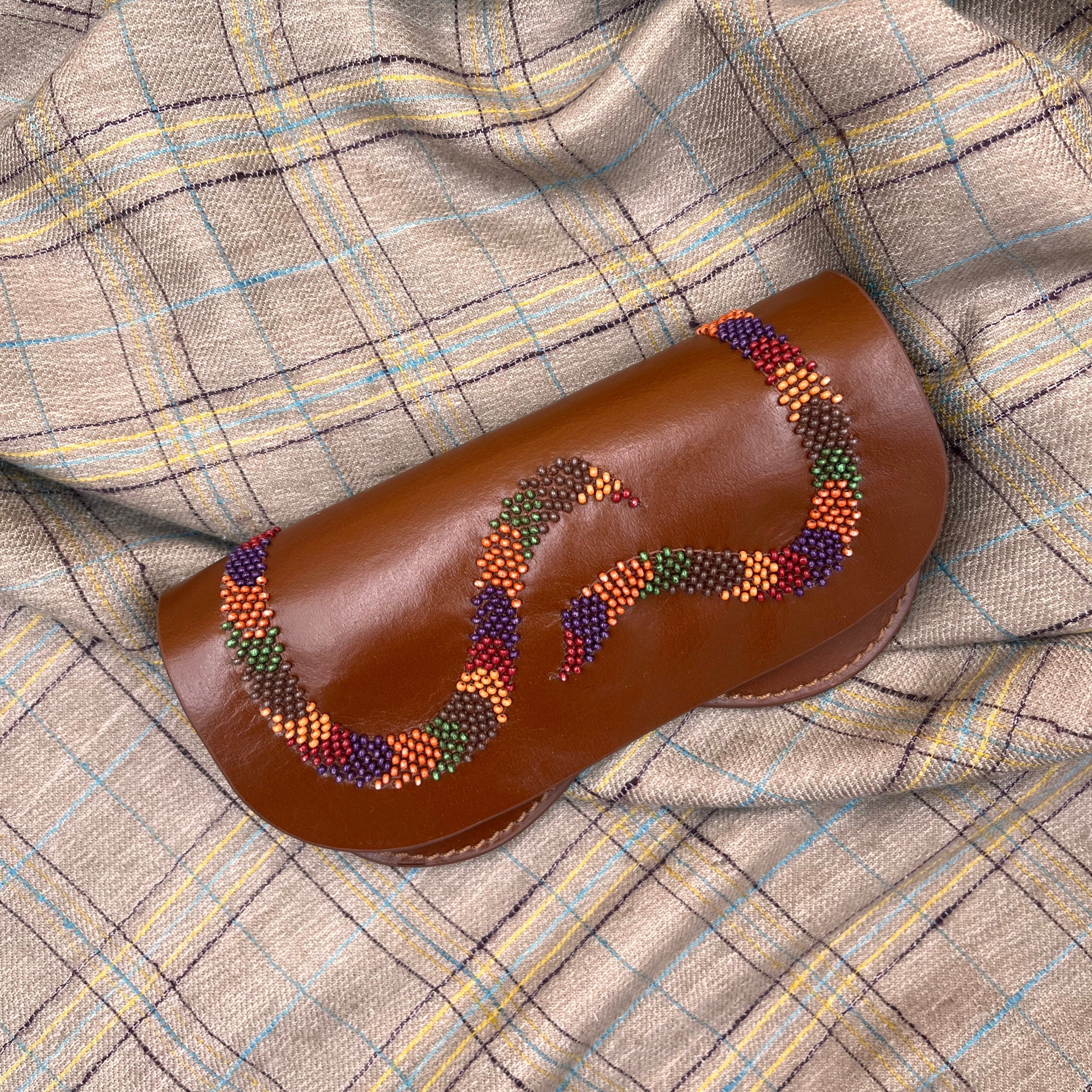 A brown leather glasses case with embroidered design of a snake, on a grey blanket. 