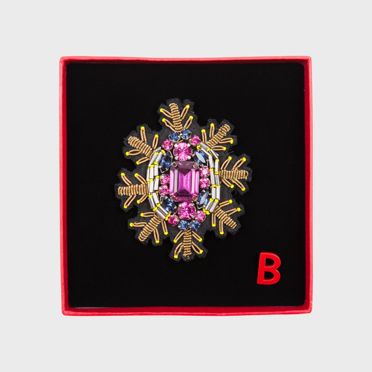 An embroidered brooch representing a snowflake with a pink stoned center in a box. 