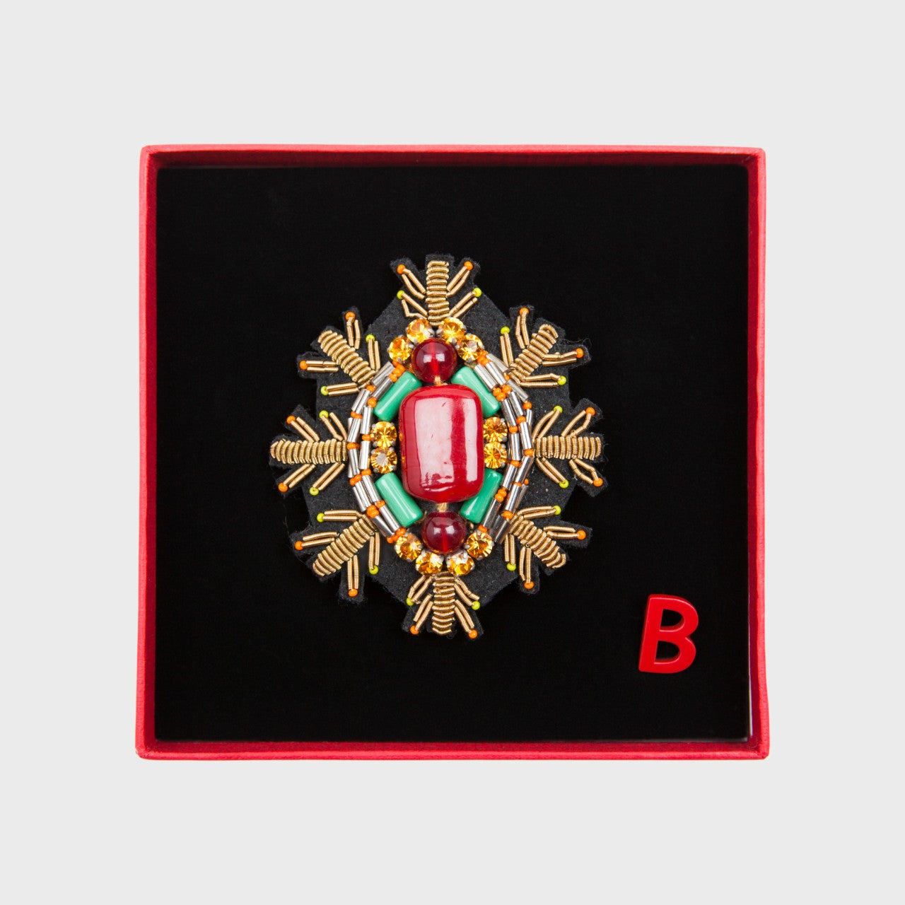 An embroidered brooch representing a snowflake with a red stoned center in a box. 
