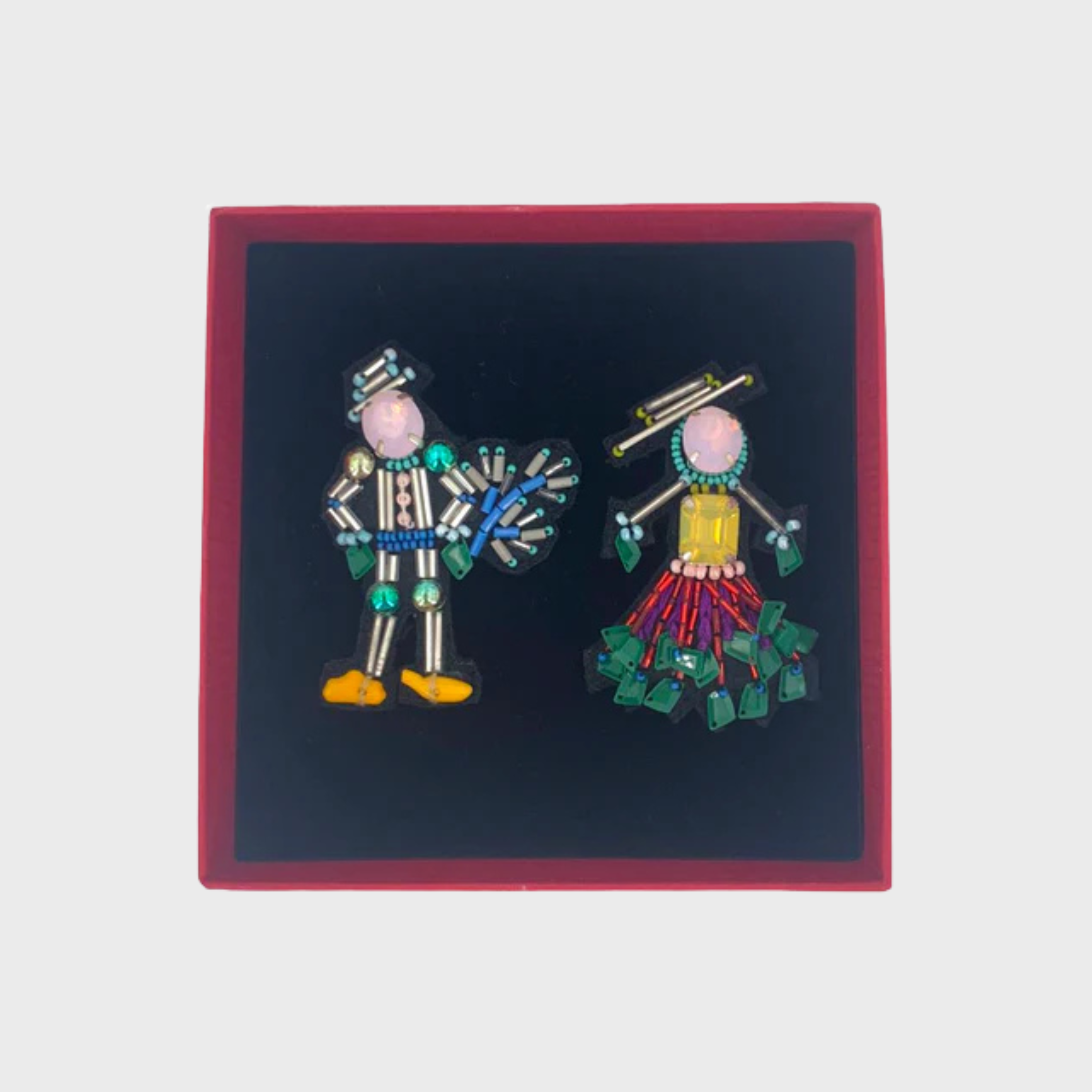 An embroidered set of two brooches representing a couple with pink heads in a box. 