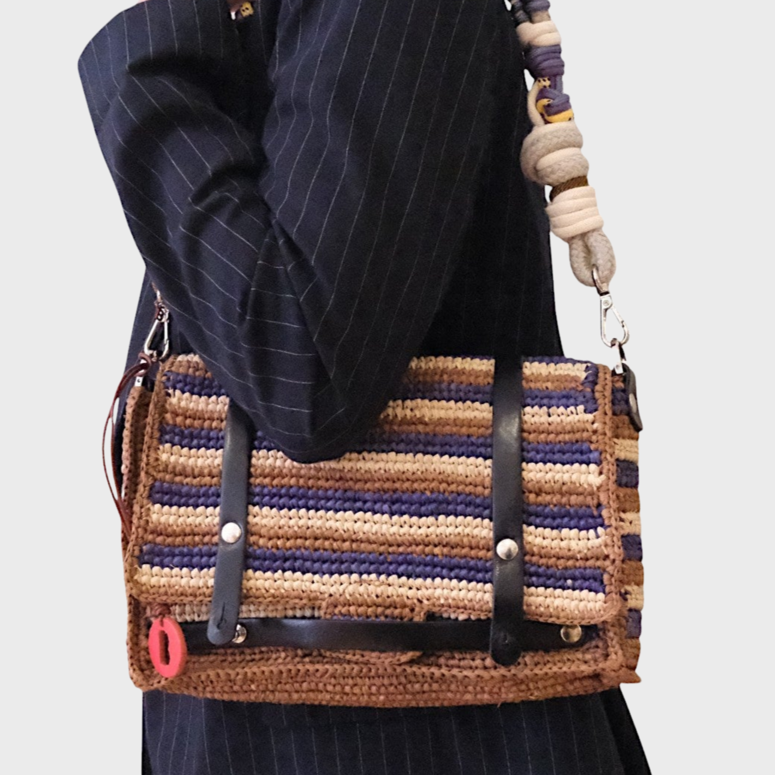 A model wearing a striped blue brown beige raffia shoulder bag with black leather straps hanging by a colorful macramé braided strap. 