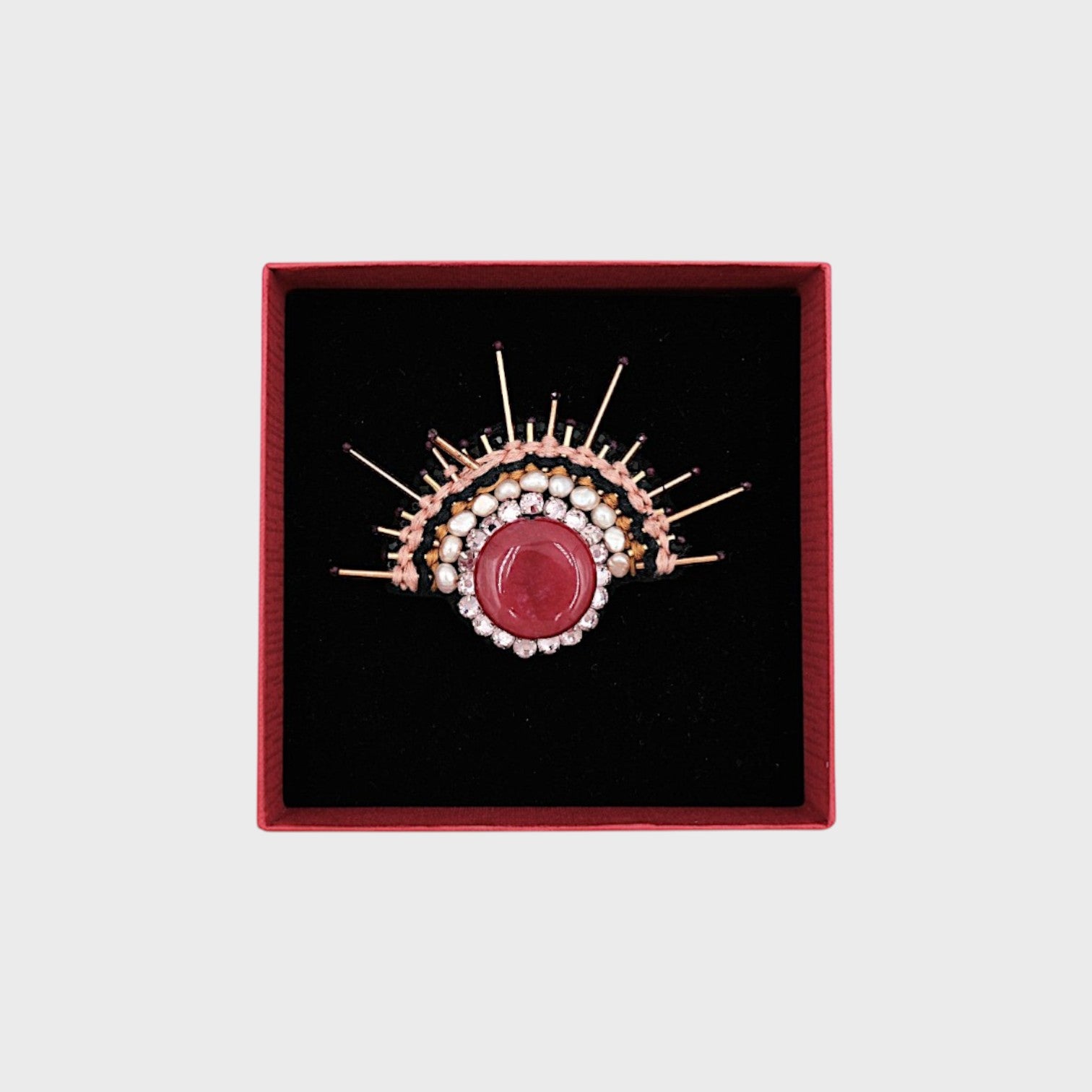 An embroidered sunrise brooch with pink details in a box. 