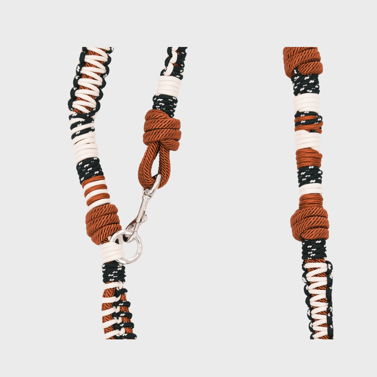 A close-up studio shot of a macramé dog leash, on a light gray background. The macramé dog leash is woven in black, white, and rust brown, with a rust brown knotted section and a silver metal clasp.
