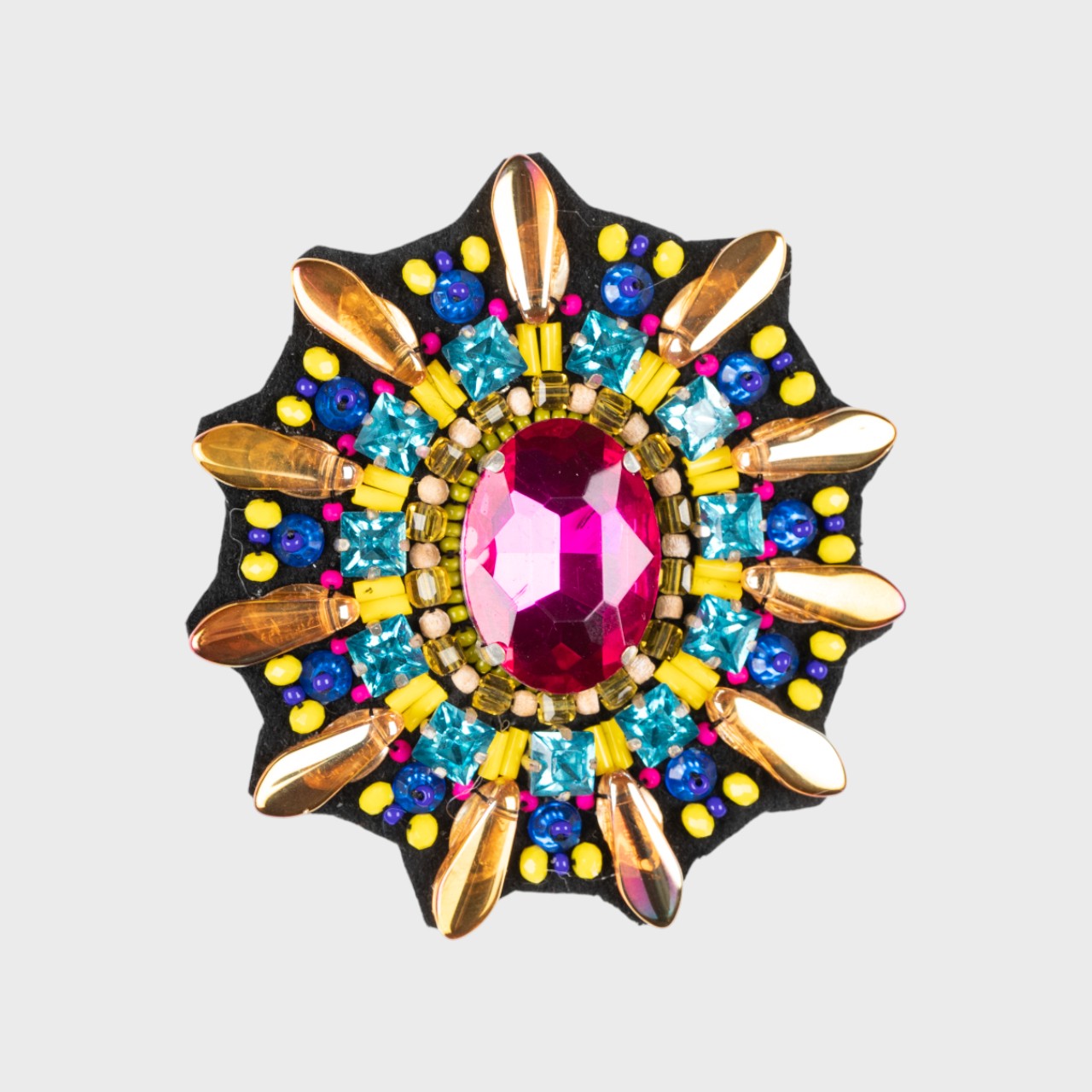 An embroidered brooch representing the shell of a turtle with a pink stone in the middle and yellow and blue details around. 