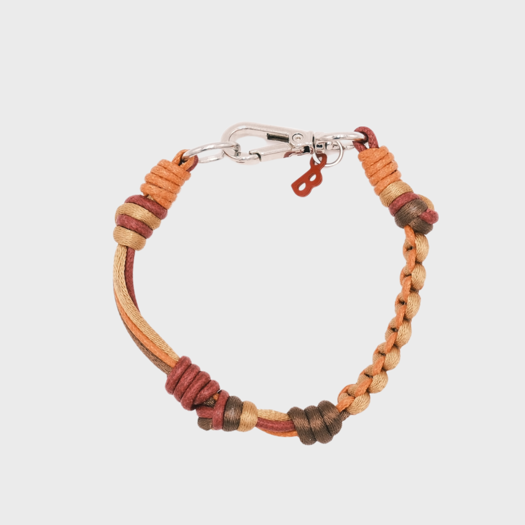 A photo taken from above of a thin braided macramé bracelet in shades of brown, orange and burgundy, with a silver clasp.