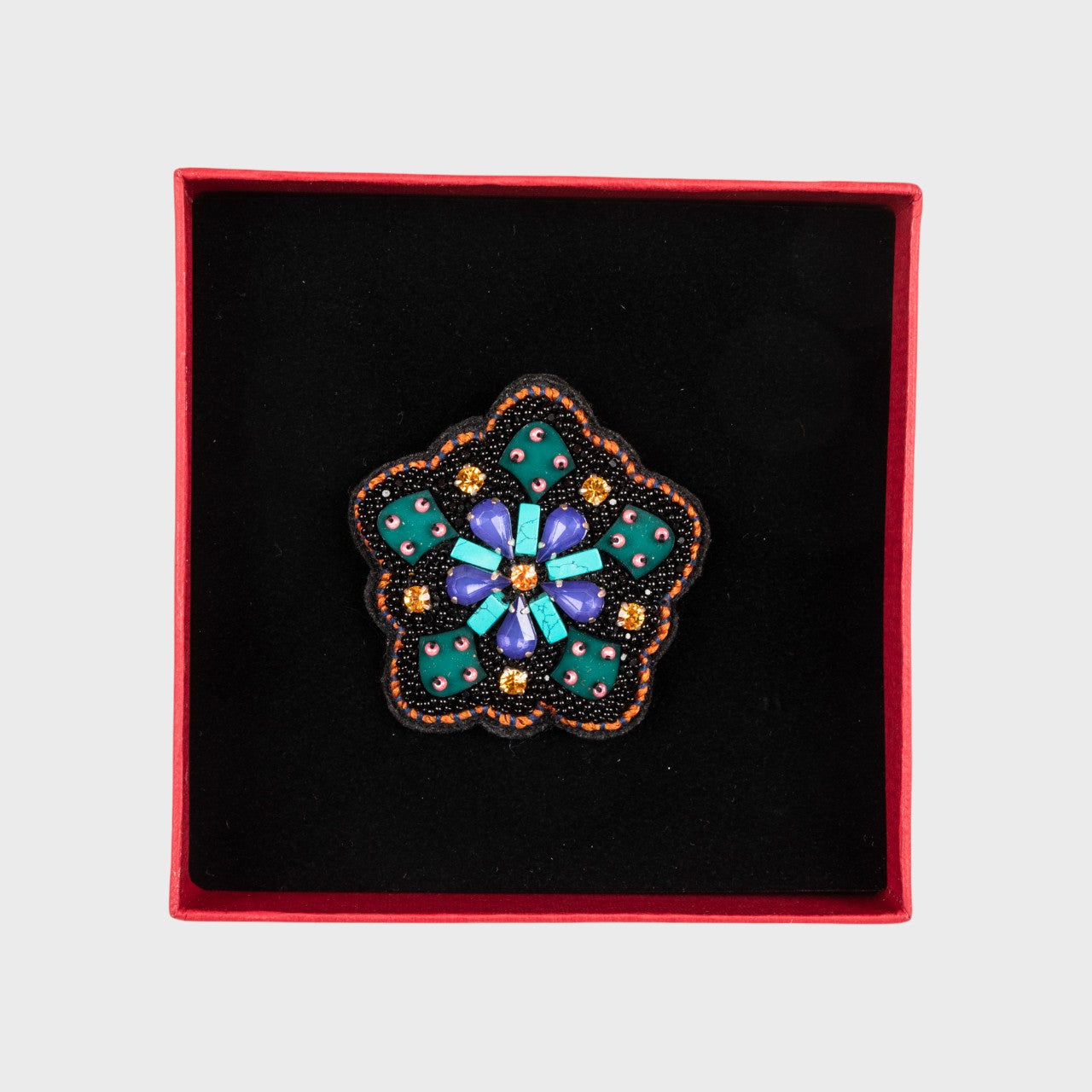 An embroidered brooch representing a wheel with green details in a box. 