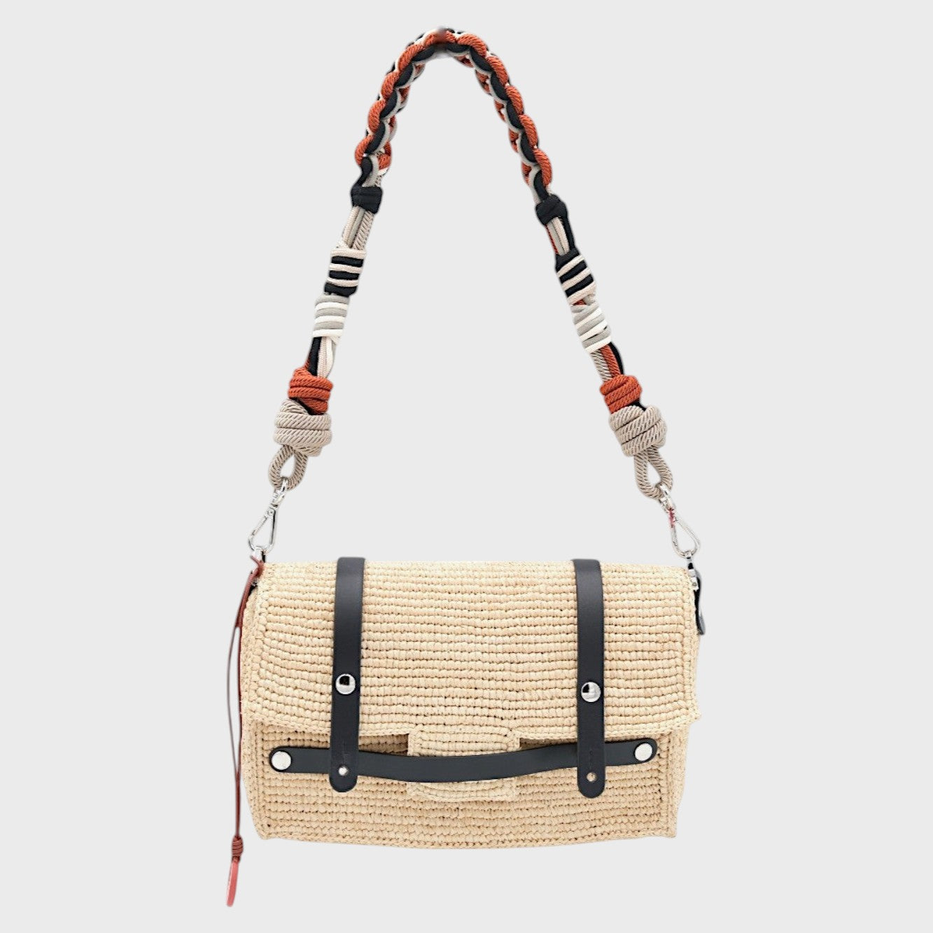 A white raffia shoulder bag with black leather straps hanging by a colorful macramé braided strap featuring grey, black and dark orange, set against a light gray background.