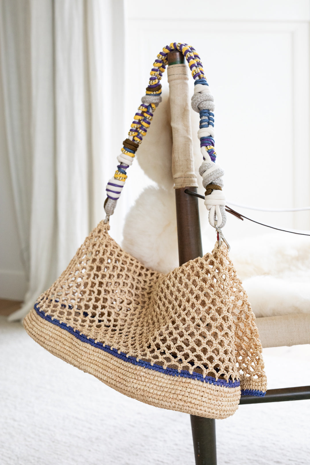 A white raffia Colin Tote Bag with blue stripes, hanging by a yellow, purple and blue beaded bag strap. 
