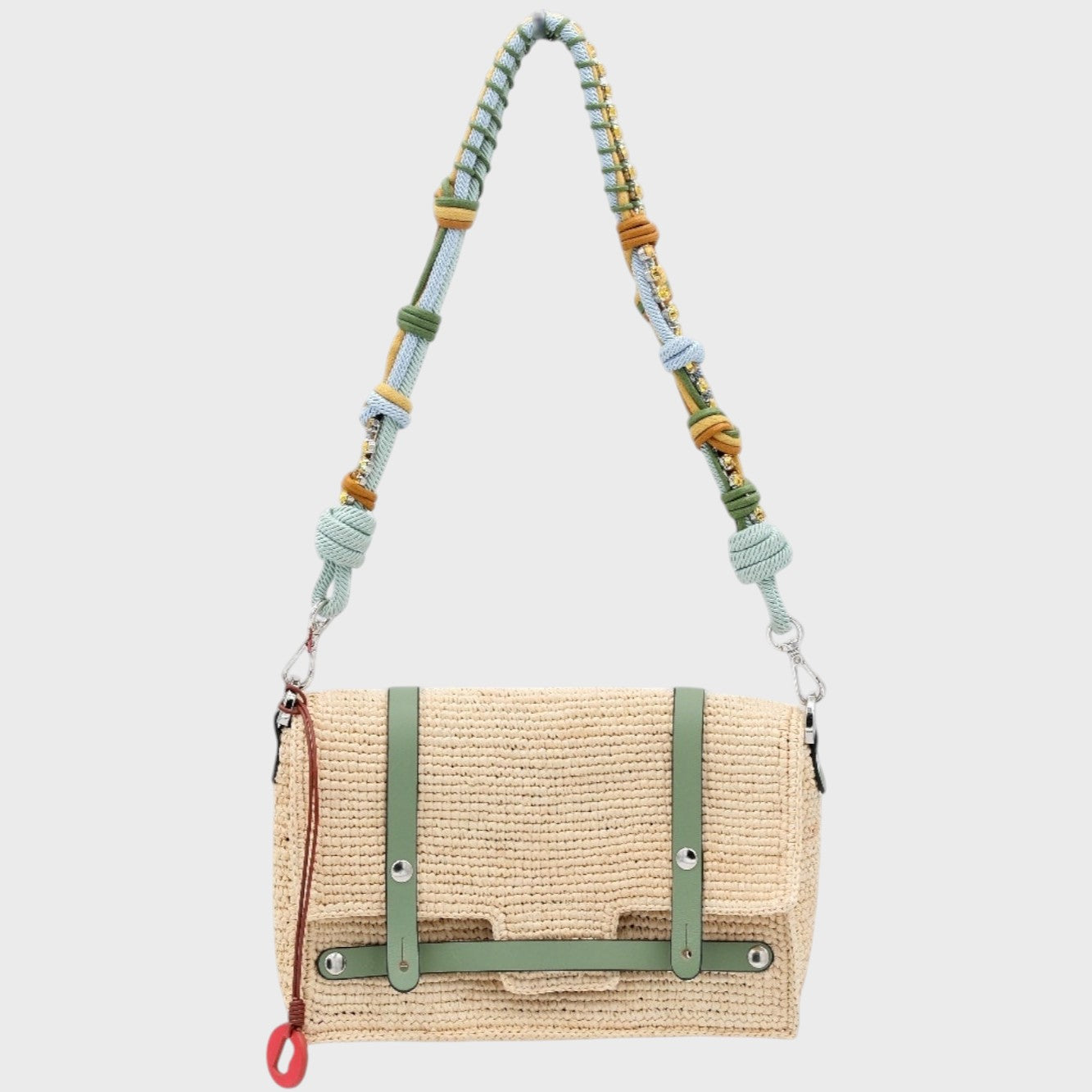 A white raffia shoulder bag with green leather straps hanging by a colorful macramé braided strap featuring light blue, green and small yellow beaded stones, set against a light gray background.
