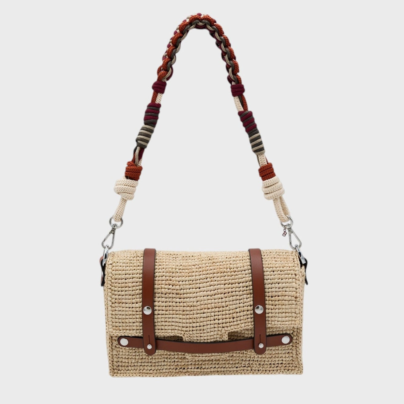 A white raffia shoulder bag with brown leather straps hanging by colorful macramé braided strap featuring white, burgundy and dark orange, set against a light gray background.