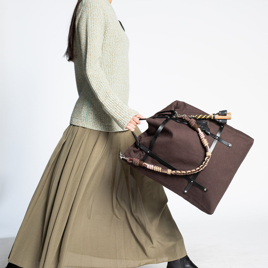 A woman in a sage green knit sweater and an olive green pleated skirt walks while carrying a brown canvas travel bag with a macramé braided strap, against a light gray studio background.