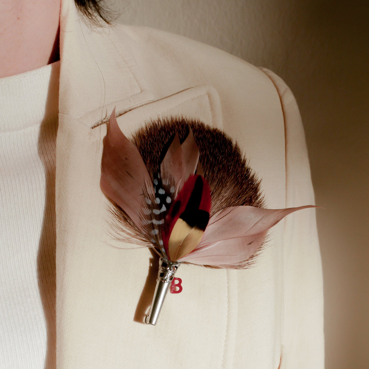 A close-up of a feather brooch with a metal pin attached, featuring a mix of brown, black, and yellow feathers, secured onto a jacket lapel.