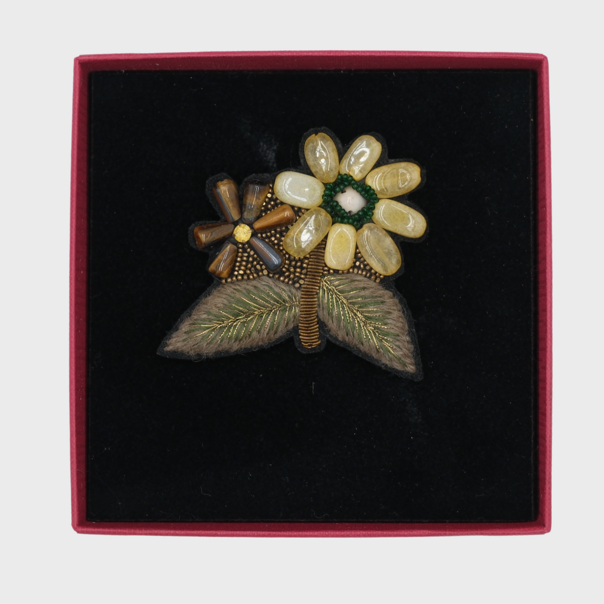 A floral brooch featuring cream and brown enamel flowers with green and white centers, set on a black velvet presentation box with a red border.