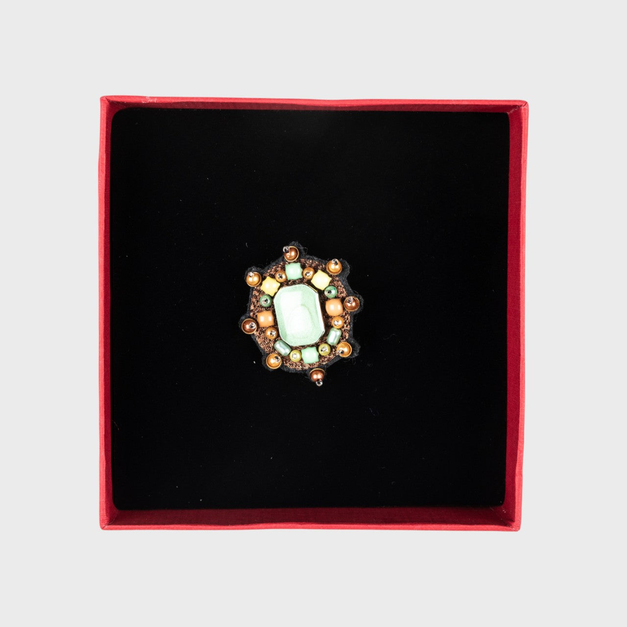 An embroidered brooch representing a small yourte with a centered green stone in a box. 