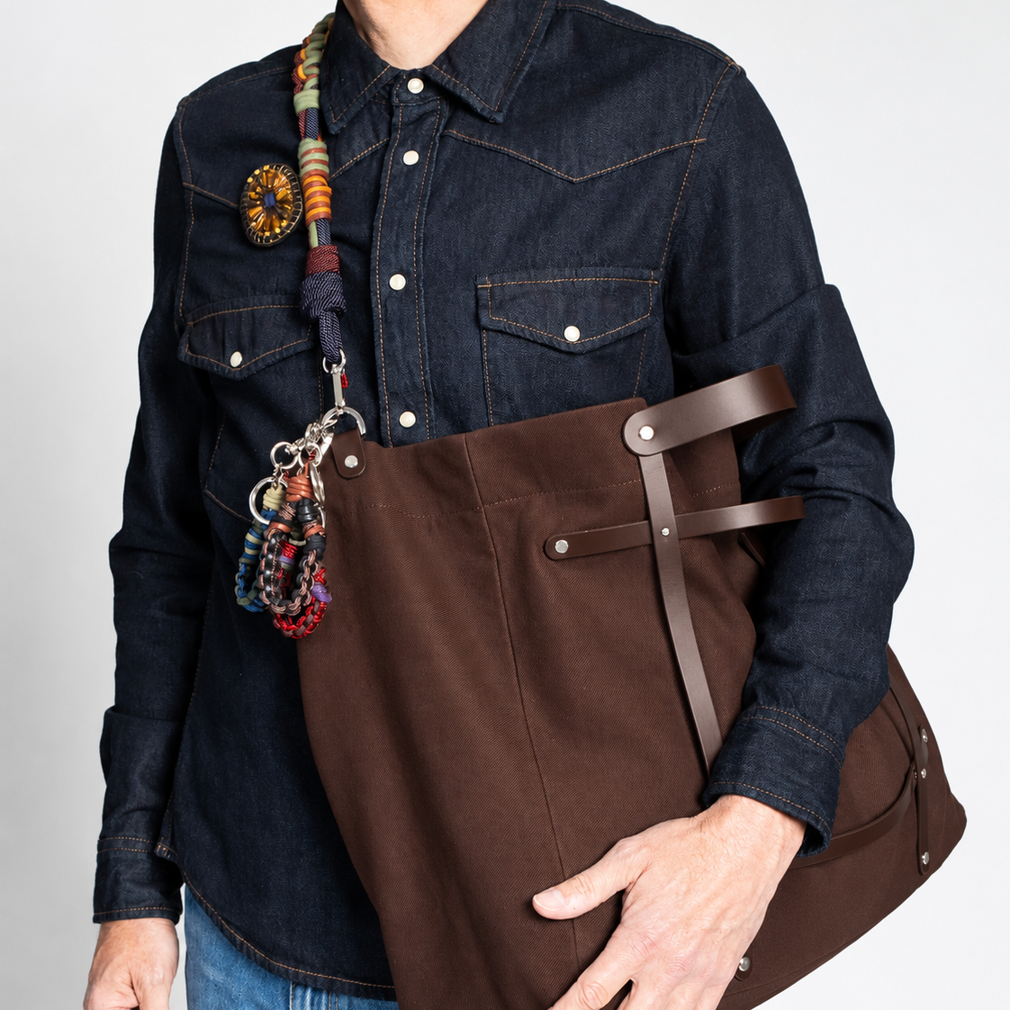 A person wearing a dark denim shirt and blue jeans carries a large brown canvas weekender bag with brown leather straps and a colorful beaded shoulder strap against a light gray background.