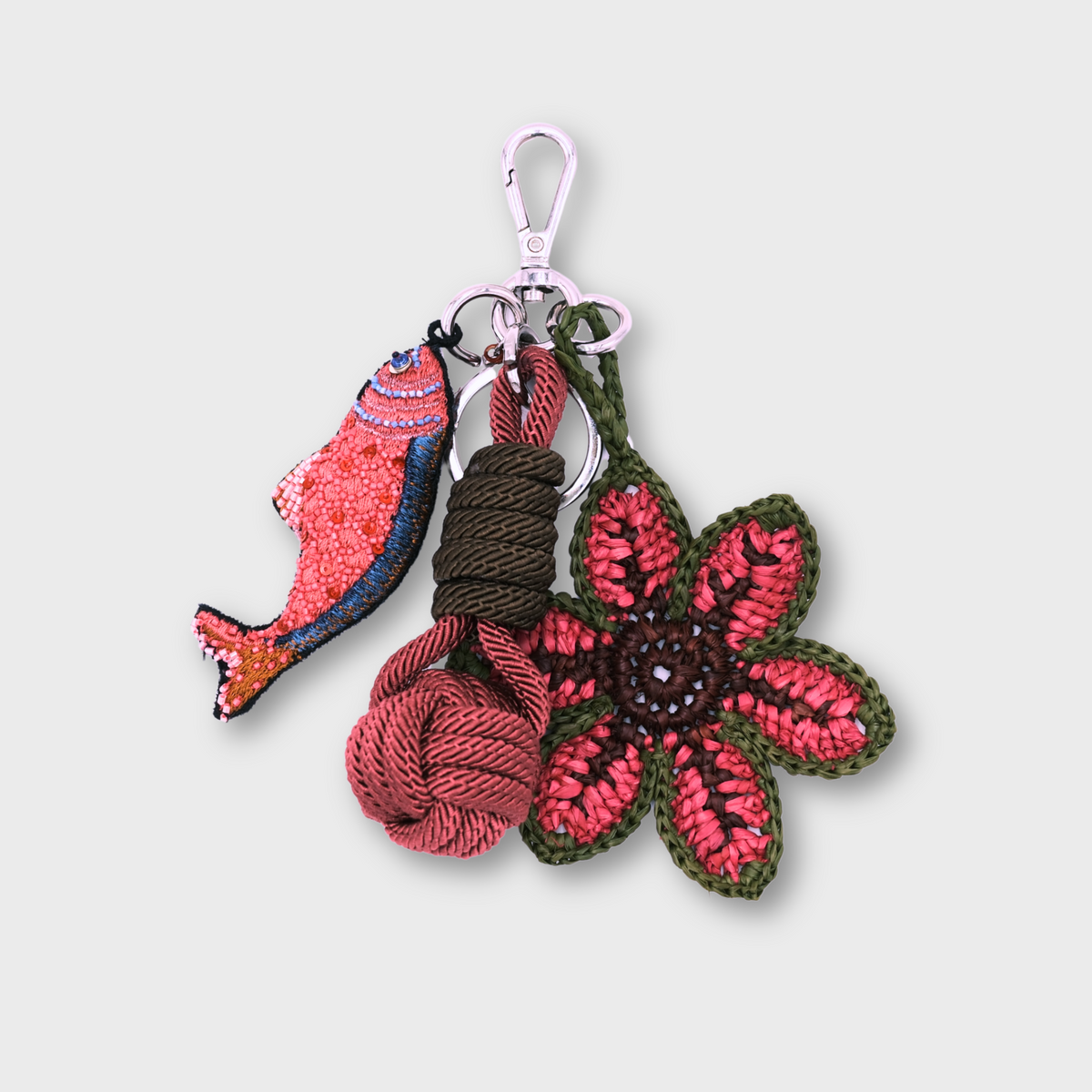A handmade macramé and raffia keychain featuring a pink and blue fish charm, a knotted prink and green ball, and a pink and green raffia flower, attached to a silver clasp.
