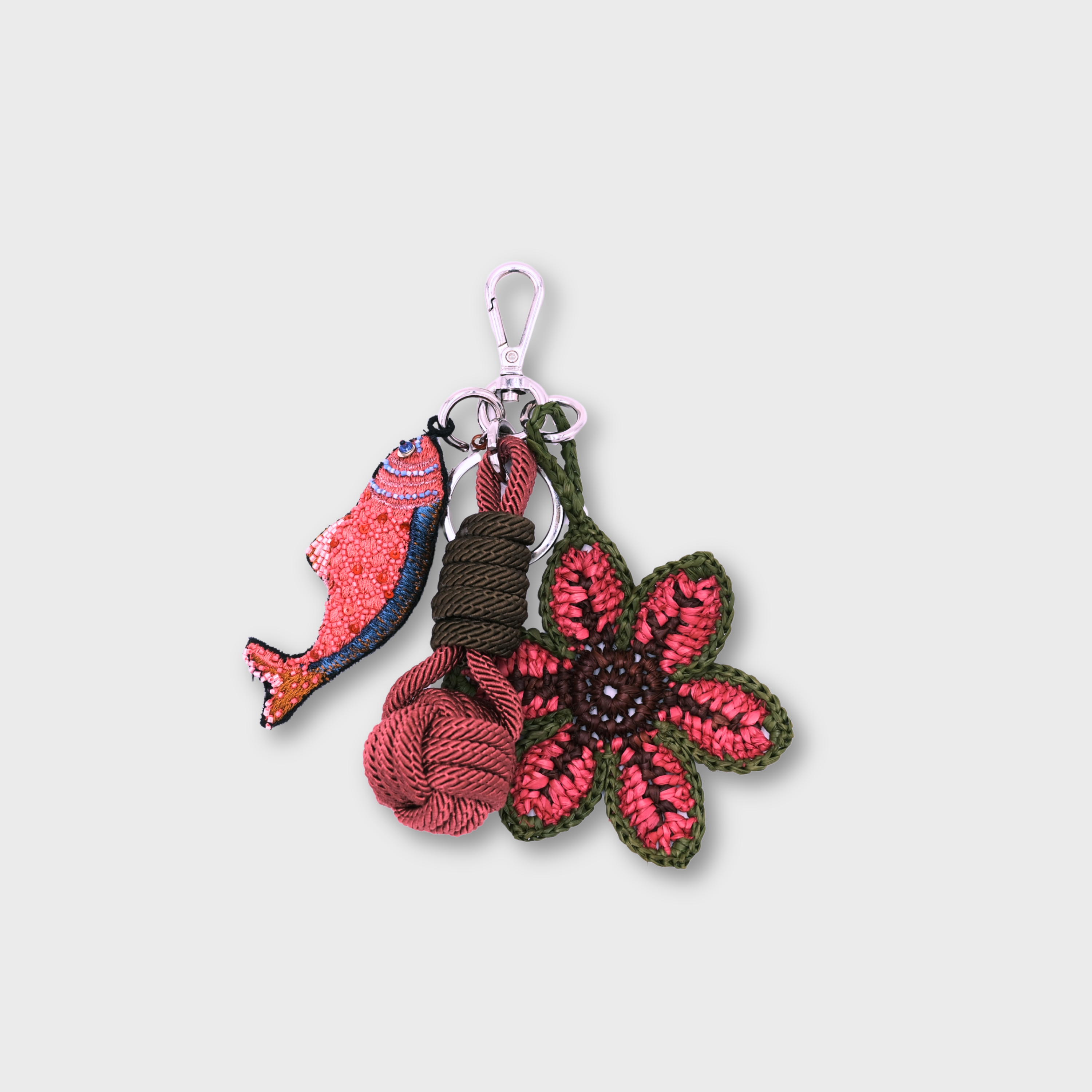 A handmade macramé and raffia keychain featuring a pink and blue fish charm, a knotted prink and green ball, and a pink and green raffia flower, attached to a silver clasp.
